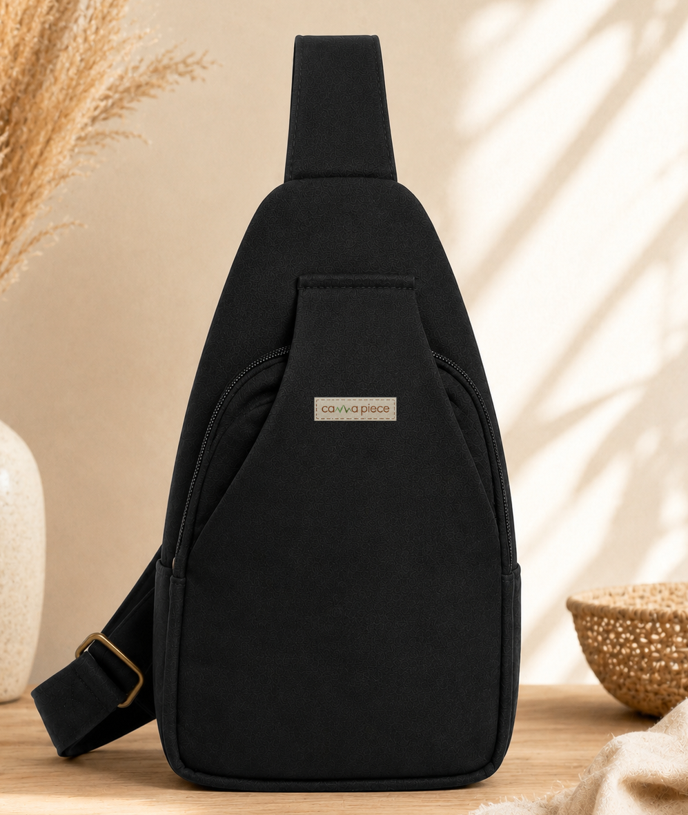 A black sling backpack with a small rectangular brand patch reading 'carvewise' is shown on a wooden surface. The scene includes a beige wall with leaf shadows, a white vase with dried grass on the left, and a woven bowl and beige cloth on the right.